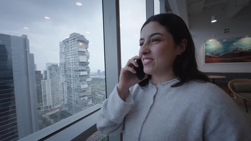 Smiling young woman enjoys a phone conversation while looking out a large window at the modern cityscape from a high-rise building