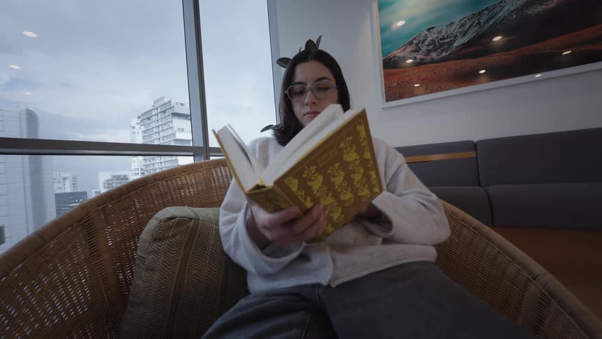 A young woman with glasses opens a book to read while sitting in a comfortable chair by a window overlooking a cityscape