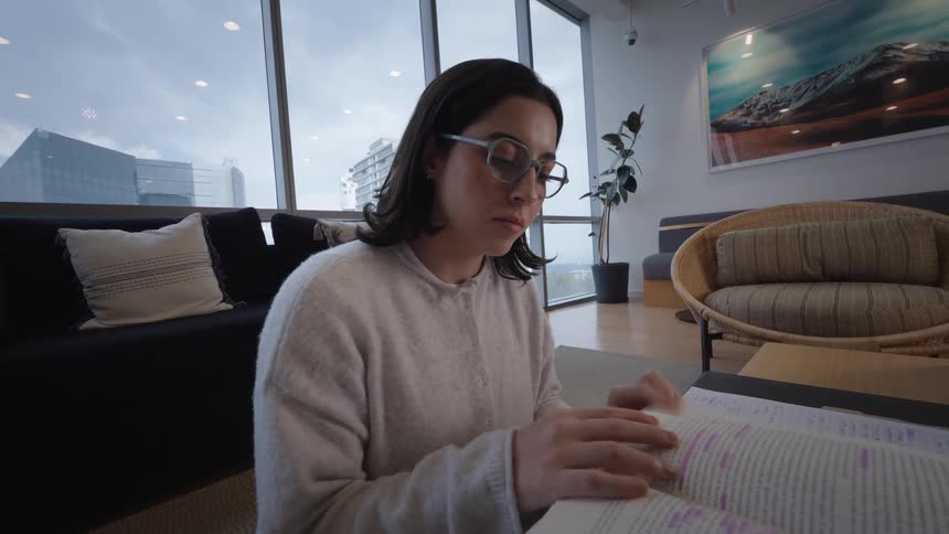 A focused young woman with glasses studies a textbook in a contemporary city apartment, slowly turning the pages