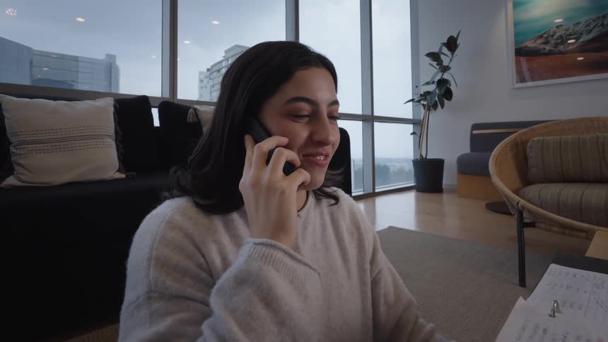 A cheerful young woman smiles while talking on her smartphone in a modern city apartment