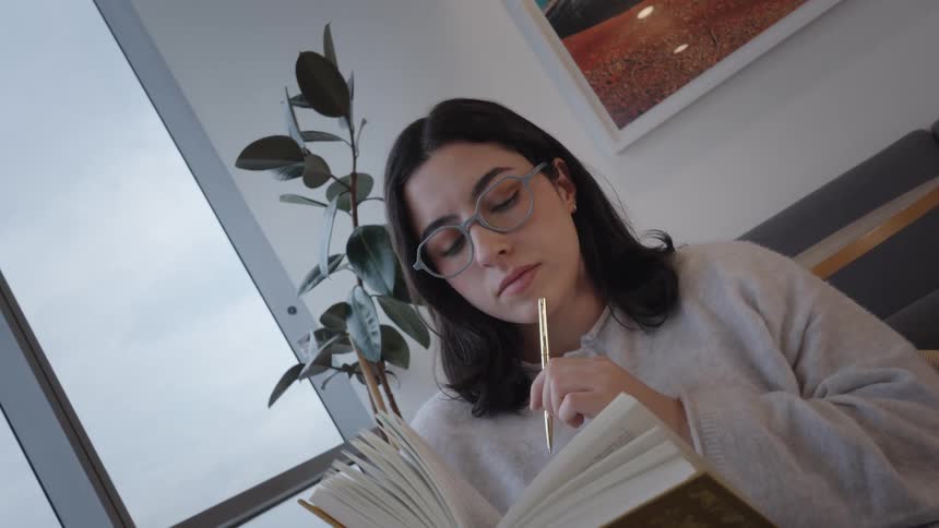 A studious young woman in glasses reads a book, then pauses thoughtfully while sitting in a modern lounge by a large window