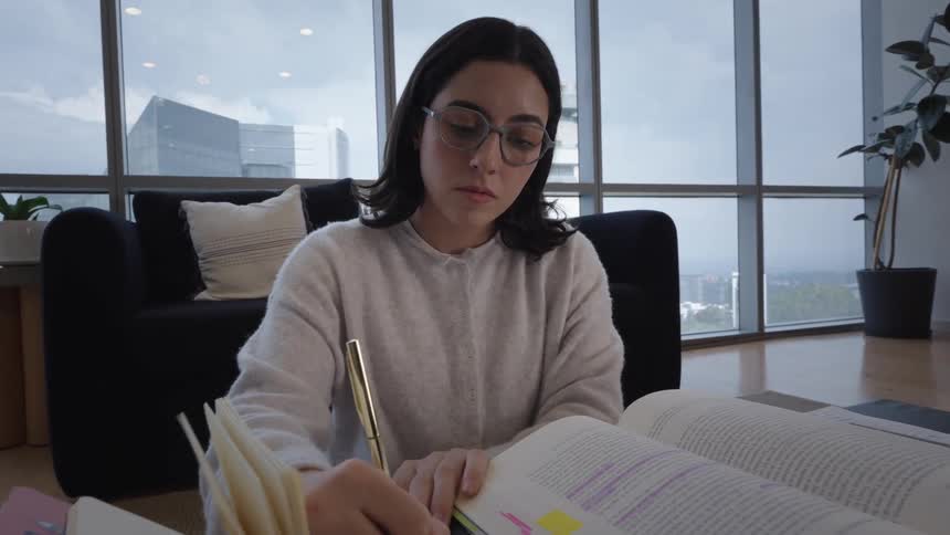 A focused young woman wearing glasses studies from highlighted textbooks and writes notes in a modern apartment with city views