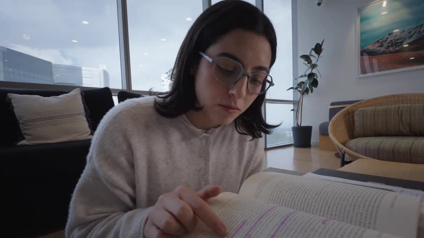 A young woman with glasses diligently studies from books in a modern apartment lounge with large windows overlooking a city