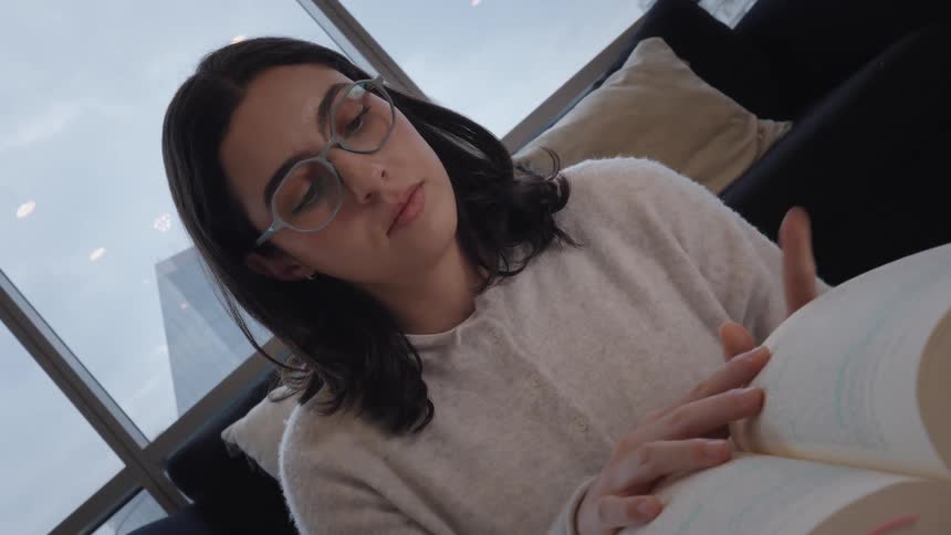 A studious young woman wearing glasses diligently reads a book, turning the pages while studying in a modern city apartment