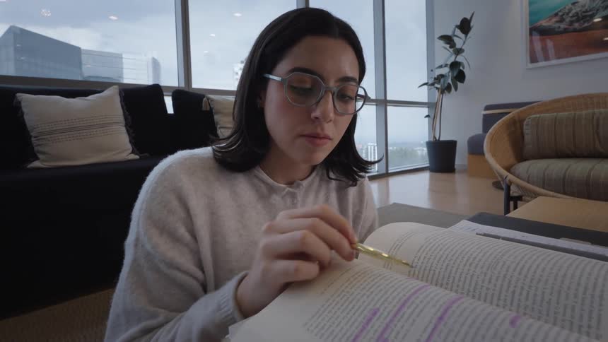 A young woman with glasses studies from a large book, surrounded by notebooks and textbooks on the floor of her modern apartment with city views
