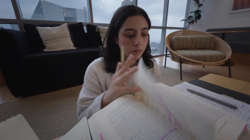 A young female student intently reads a highlighted textbook on the floor of her modern apartment