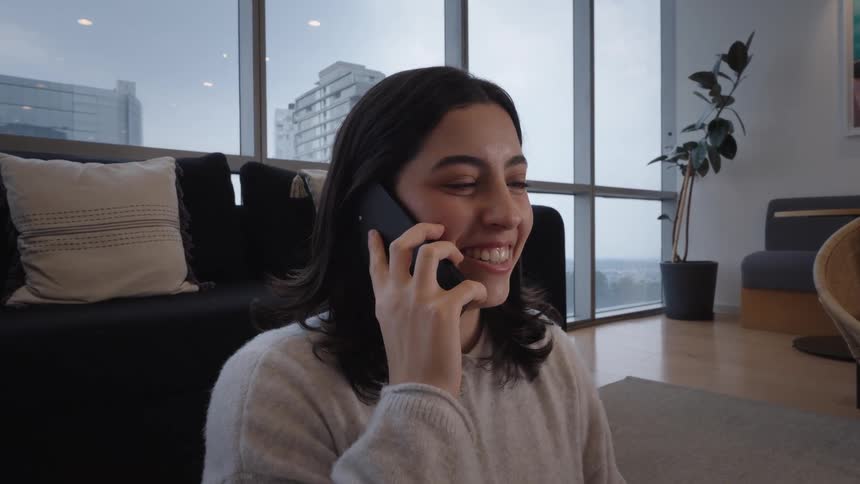 A smiling young woman sits on the floor of her modern apartment and has a pleasant conversation on her smartphone