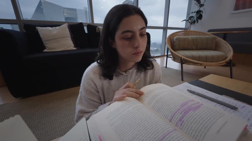 A focused female student diligently reads a highlighted textbook in her modern city apartment