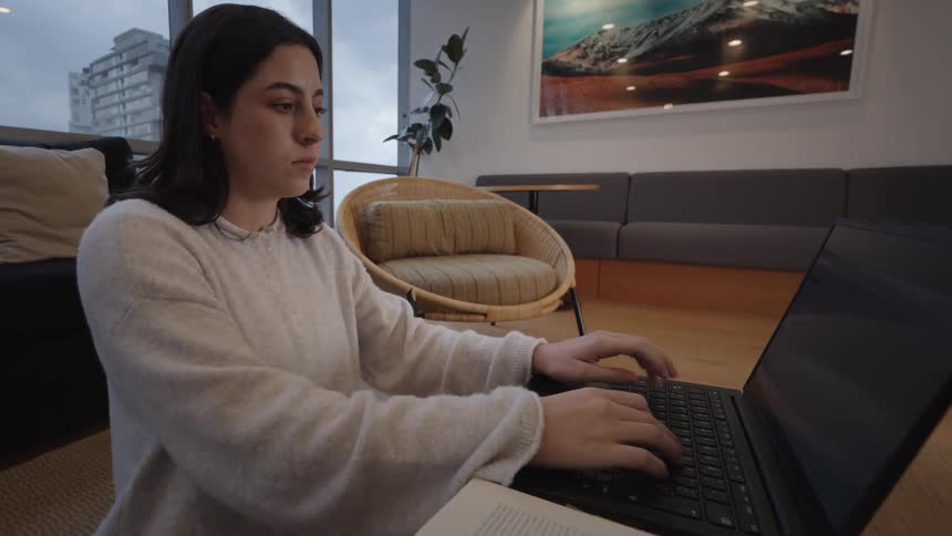 A young female student sits on the floor of a modern living room, reading a book and typing on a laptop computer
