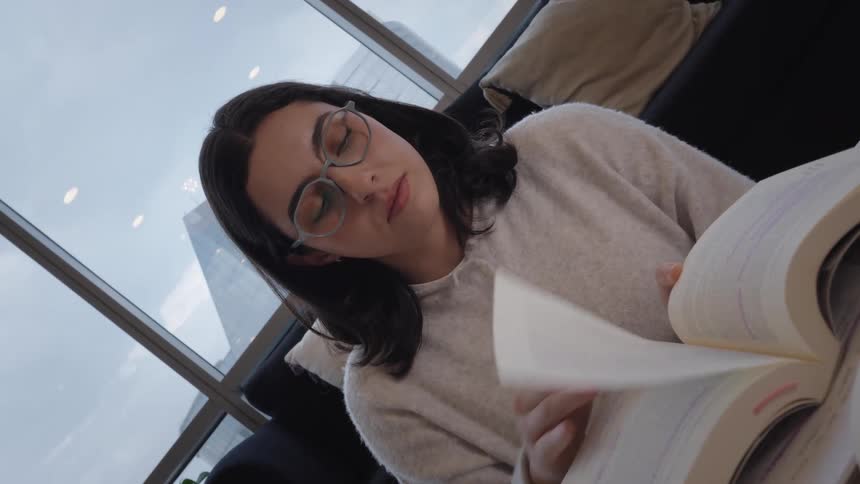 A studious young woman puts on her glasses and begins to read a book in a modern apartment with a view of the city skyline