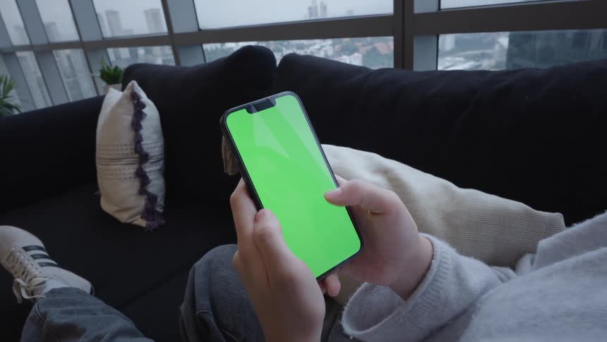 A woman relaxes on a couch in her city apartment while scrolling through her smartphone with a green screen
