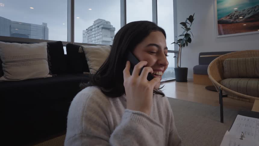 A young woman enjoys a pleasant phone conversation, smiling and laughing while sitting on the floor of her modern apartment