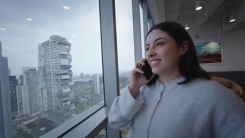 A young woman in a high-rise building answers a call on her smartphone while looking out the window at the cityscape