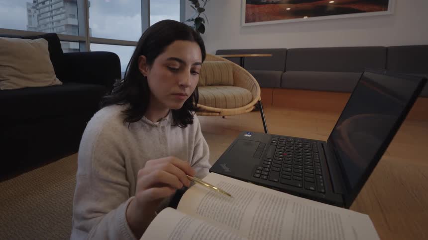 A young female student reads from a textbook and then types on her laptop while studying on the floor of a modern living room