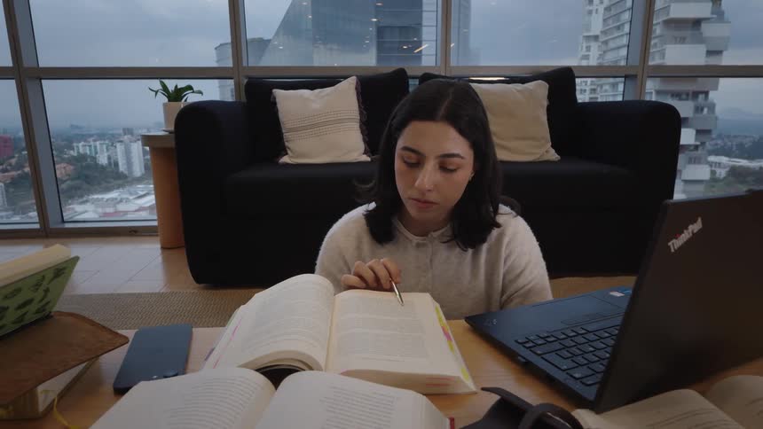 A young woman studies intently from a textbook at a low table in her modern city apartment
