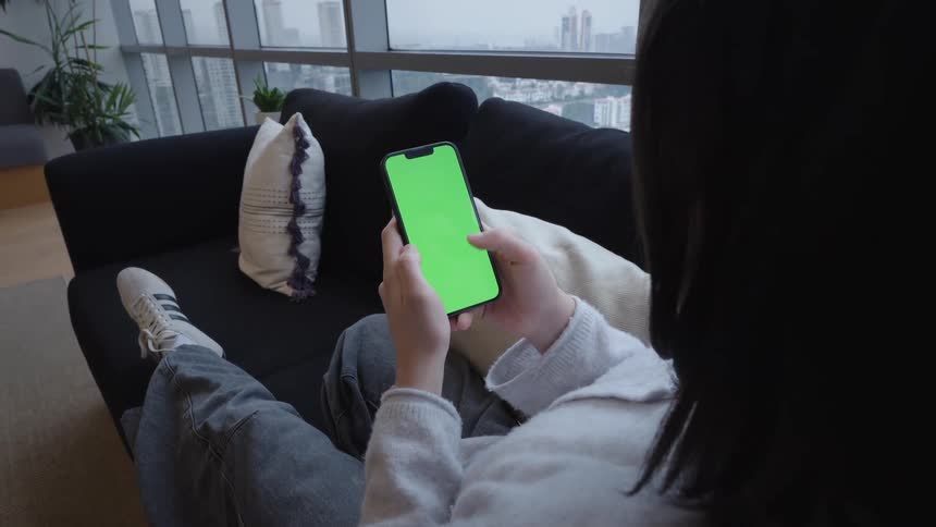 Over-the-shoulder view of a woman lounging on a sofa and using a smartphone with a chroma key green screen in front of a large window with a city view
