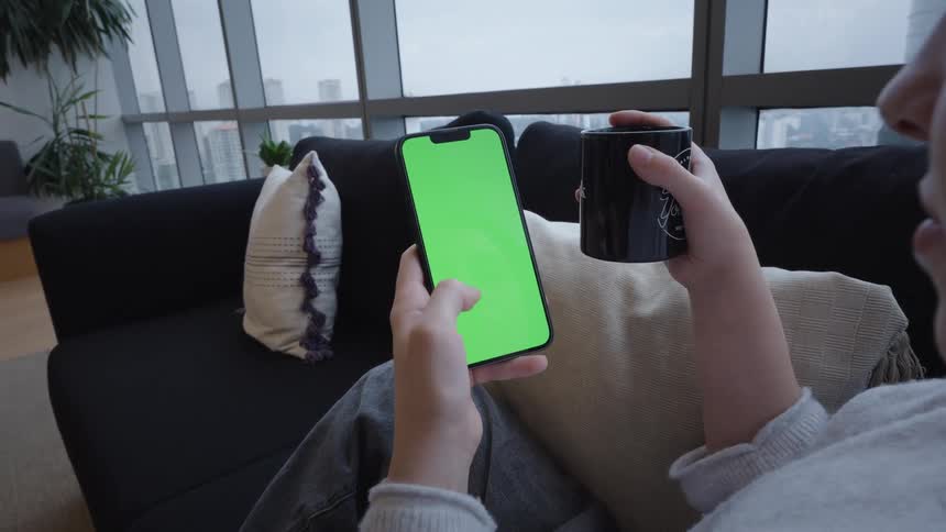 A woman relaxes on a sofa in her modern high-rise apartment, holding a mug and scrolling on her smartphone with a green screen