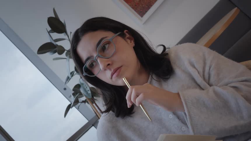 Thoughtful young woman in glasses reads a book and contemplates while sitting in a cozy chair by a city-view window