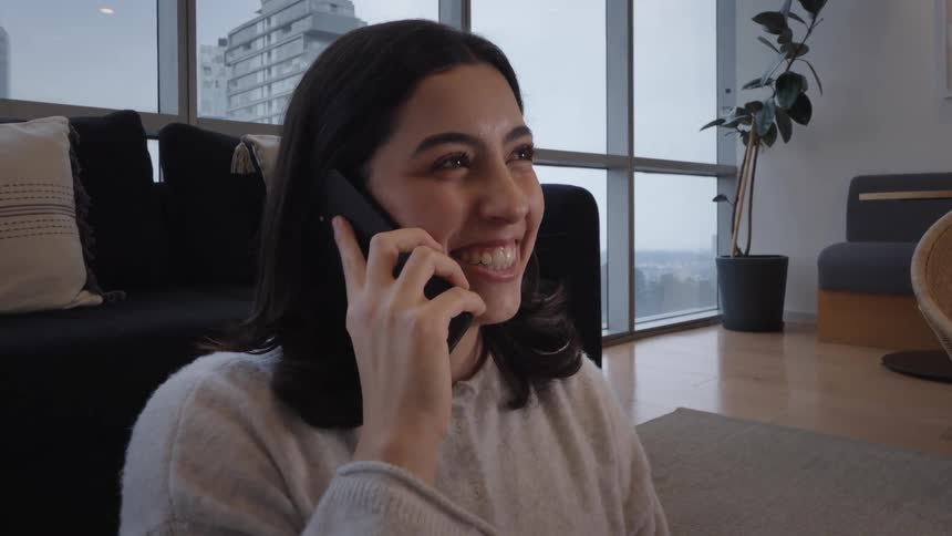 A cheerful young woman smiles and laughs while talking on her phone in a modern city apartment