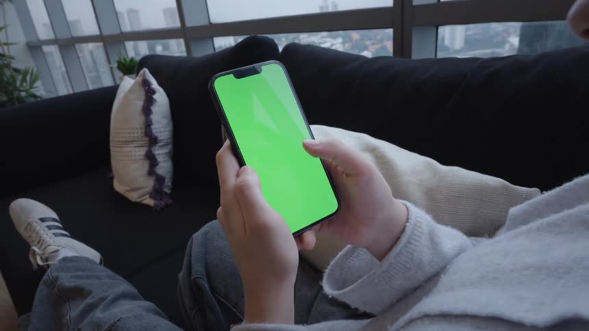 Over-the-shoulder shot of a woman relaxing on a sofa and scrolling through her smartphone with a green screen, with a cityscape visible through the window