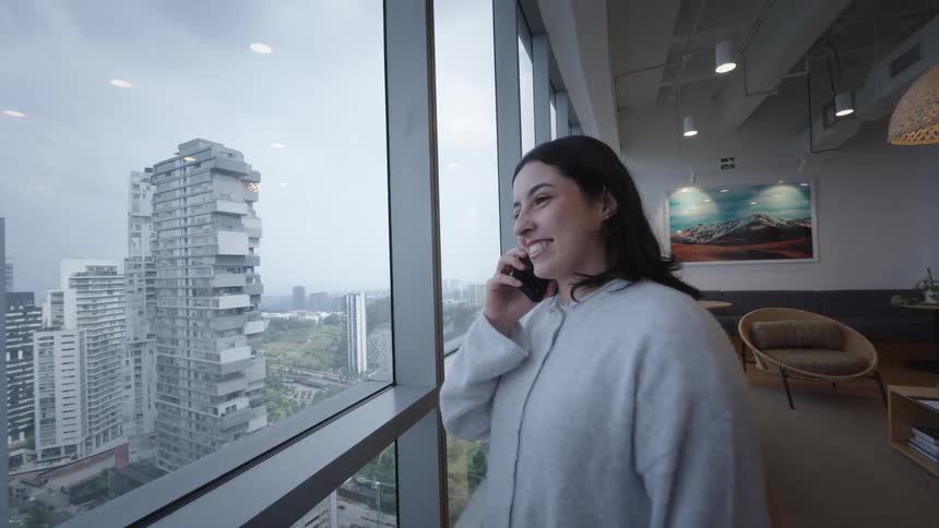 A smiling young woman talks on the phone while standing by a large window with a view of a modern city skyline