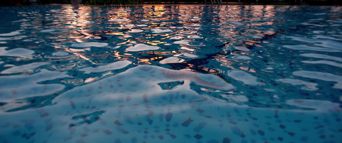 The tranquil surface of a swimming pool ripples gently, reflecting the warm colors of a beautiful sunset