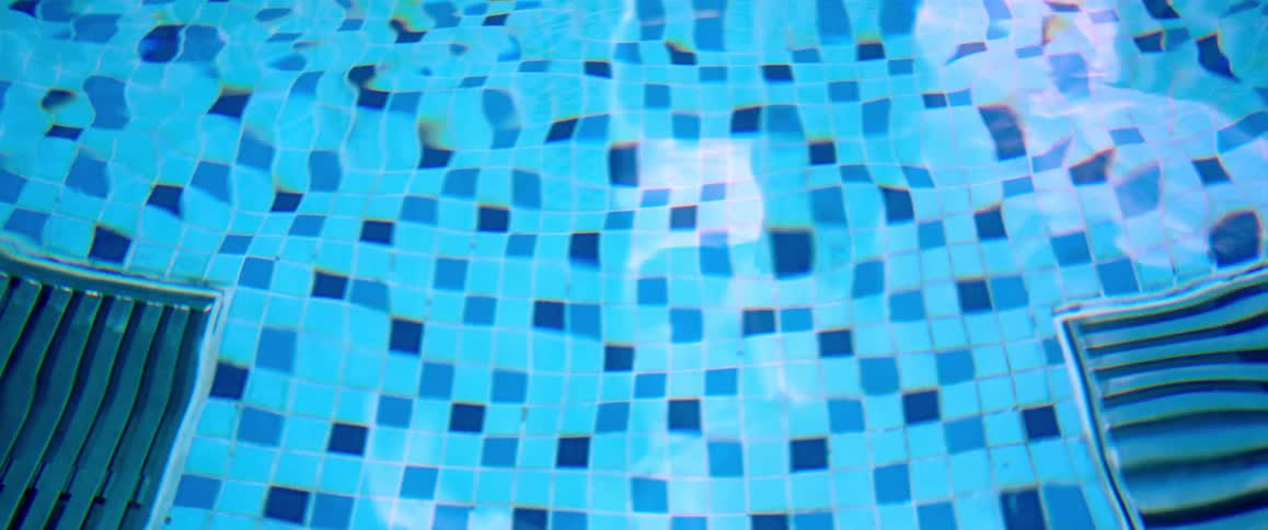 Gentle ripples move across the clear blue water of a tiled swimming pool, with sunlight reflecting on the surface