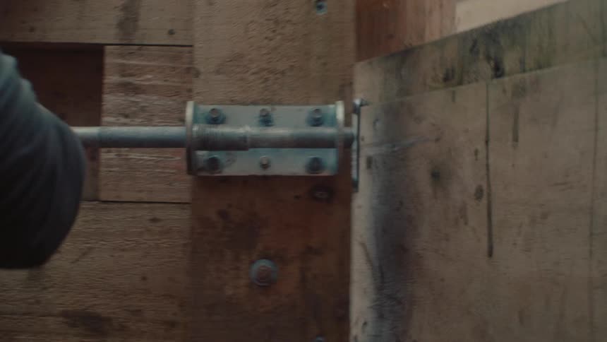 A close-up shot shows a hand sliding a metal spring-loaded bolt to lock a rustic wooden gate