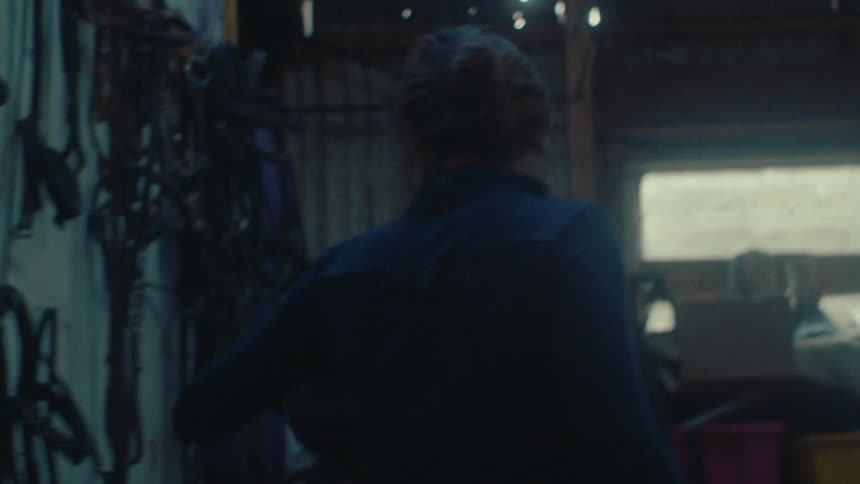 A woman grabs her riding helmet from a tack room wall in a dimly lit stable