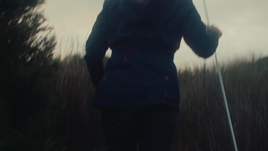 A woman in a blue jacket with a walking stick makes her way up a grassy hill on an overcast day