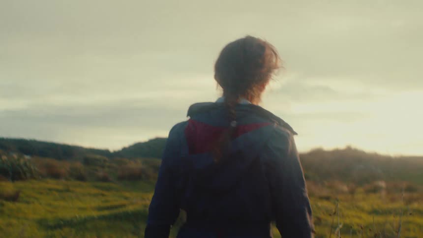 A slow-motion tracking shot follows a woman with braided hair as she walks through a golden, sun-flared field at sunset