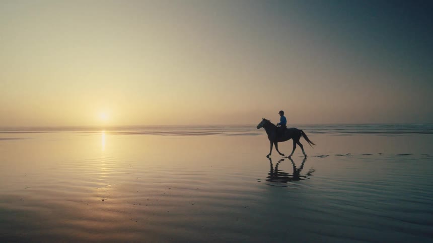 A person rides a horse through the shallow reflective waters on a beach at sunrise