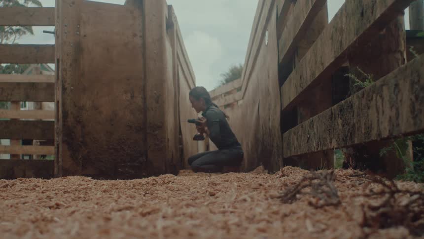 A woman kneels on wood shavings inside a narrow wooden enclosure and uses a power drill to make repairs