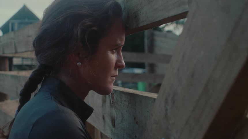 A sad and concerned woman leans her head against a wooden fence on a farm at dusk