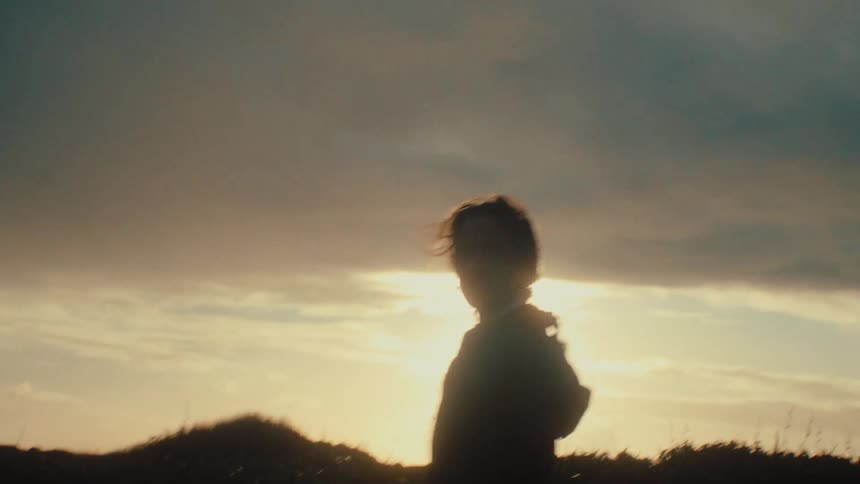 A silhouetted woman walks through a grassy field in slow motion as the golden sun sets behind her