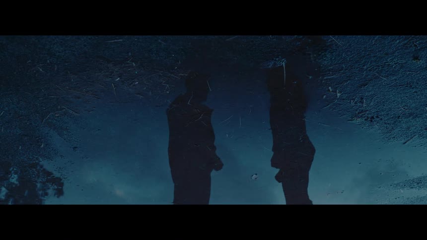 A cinematic shot shows the mysterious reflection of two silhouetted figures standing together in a dark puddle at night