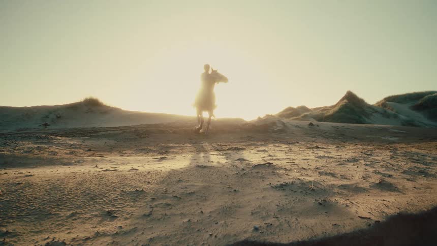 A lone rider on horseback slowly approaches through sandy dunes, silhouetted by the brilliant setting sun in a cinematic slow-motion shot