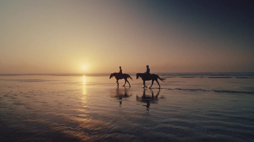 A serene silhouette of two people horseback riding on a wet sandy beach as the sun sets over the ocean