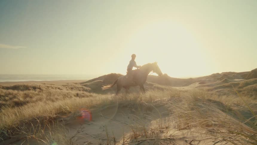 A woman gracefully rides her horse in slow motion through sandy dunes along the coast during a beautiful golden hour
