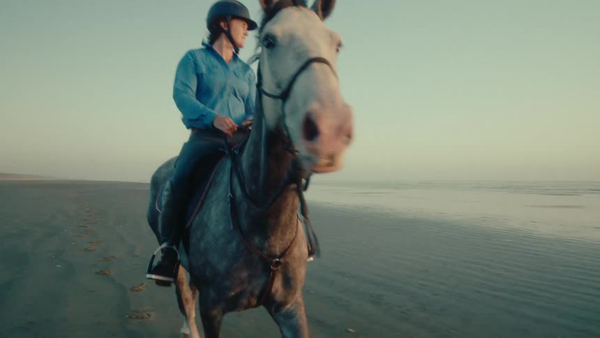 A woman wearing a helmet rides a dapple gray horse in slow motion along a wide sandy beach next to the ocean at sunrise