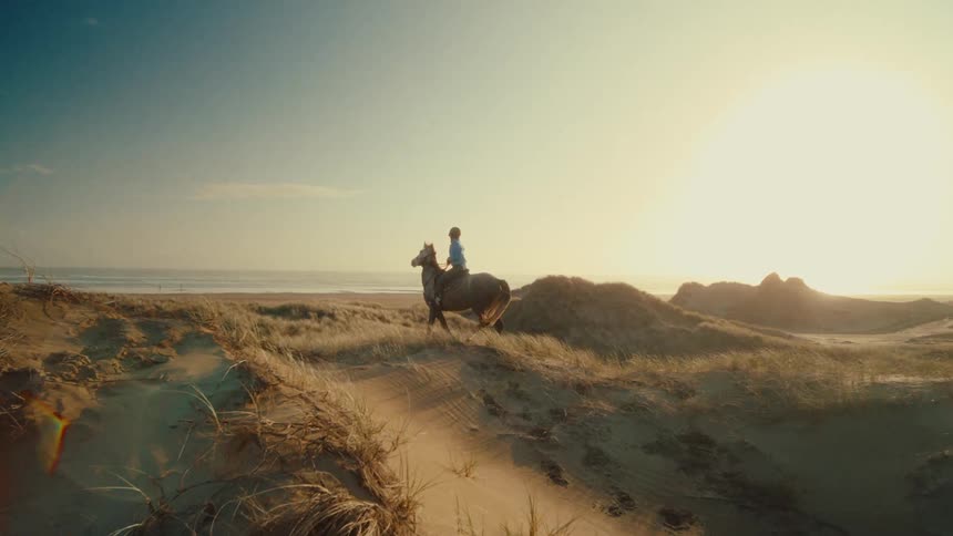A person riding a horse travels through sandy dunes towards the ocean at sunset