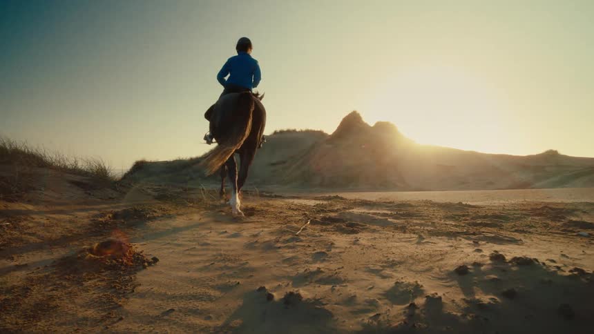 An equestrian rides a powerful horse through vast sand dunes, walking away into the golden sunset in slow motion