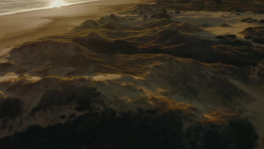 A slow aerial push shot over rolling, grassy sand dunes next to the beach during a golden sunset