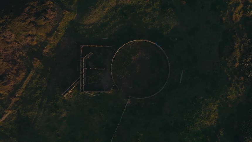 A static top-down aerial view of an empty circular livestock pen on a grassy farm during the golden light of sunrise