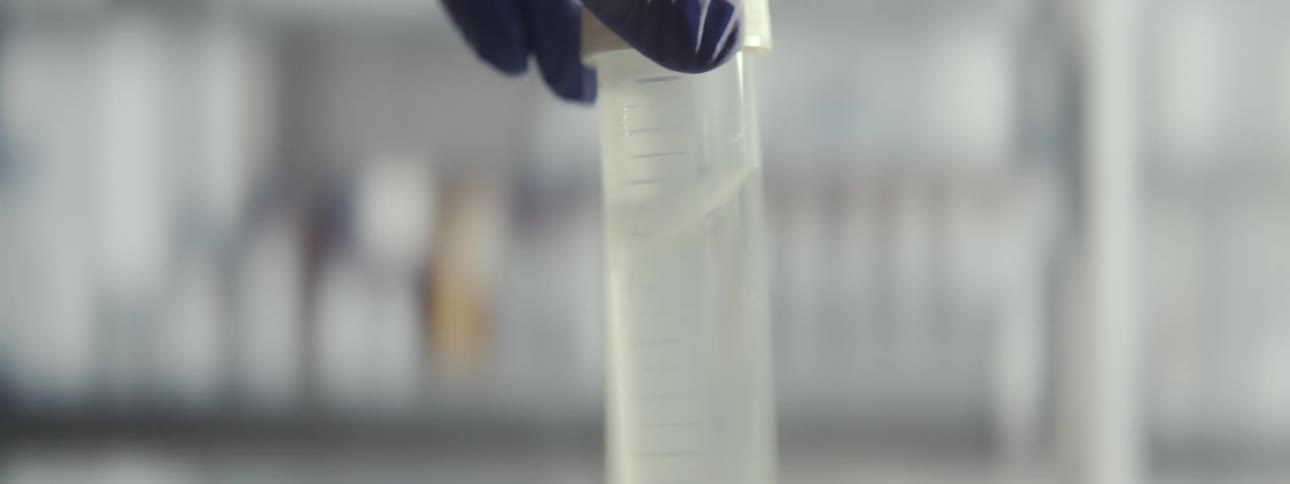 A scientist's gloved hand swirls a clear liquid containing a white substance in a test tube inside a laboratory