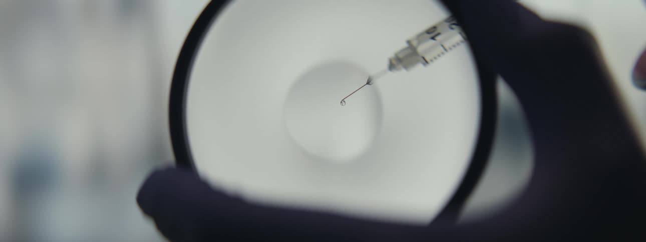 Close up of a syringe needle with a drop of liquid being examined through a magnifying glass in a laboratory
