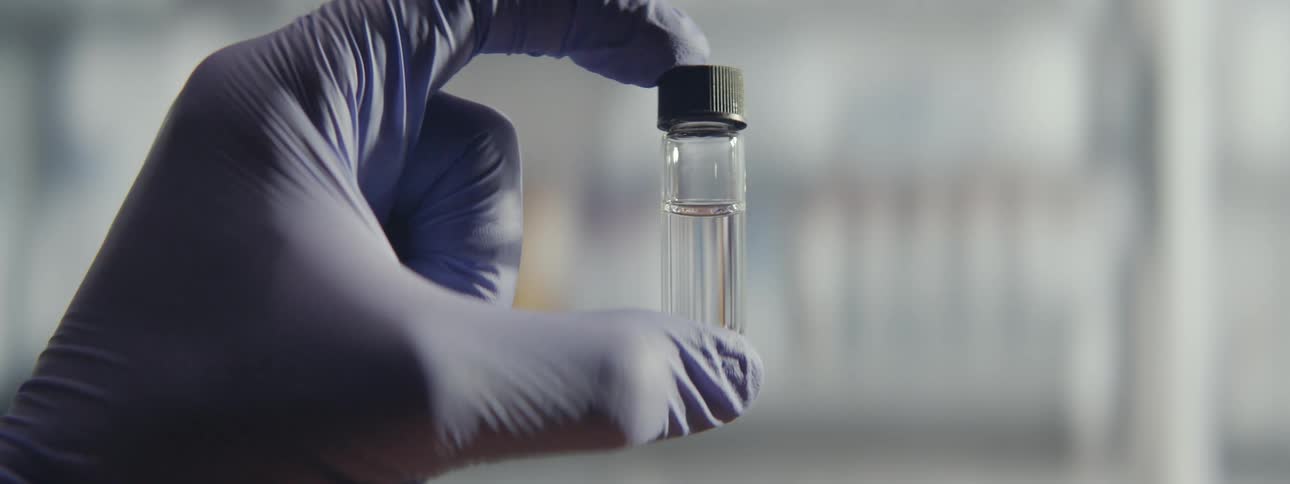 Close up of a scientist's gloved hand holding a sealed glass vial of clear liquid in a laboratory