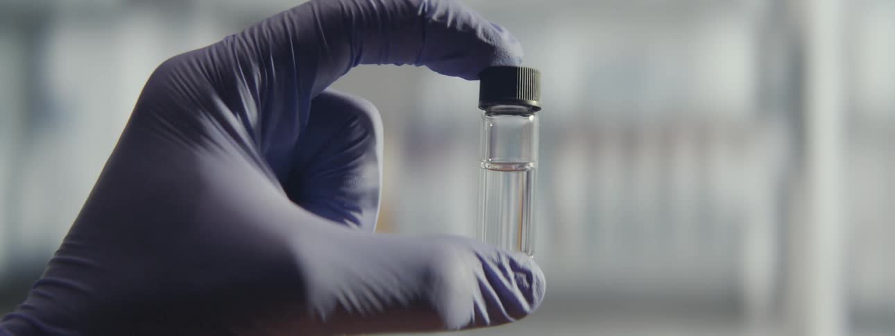 A gloved hand holds up a small glass vial filled with a clear liquid for inspection in a laboratory setting