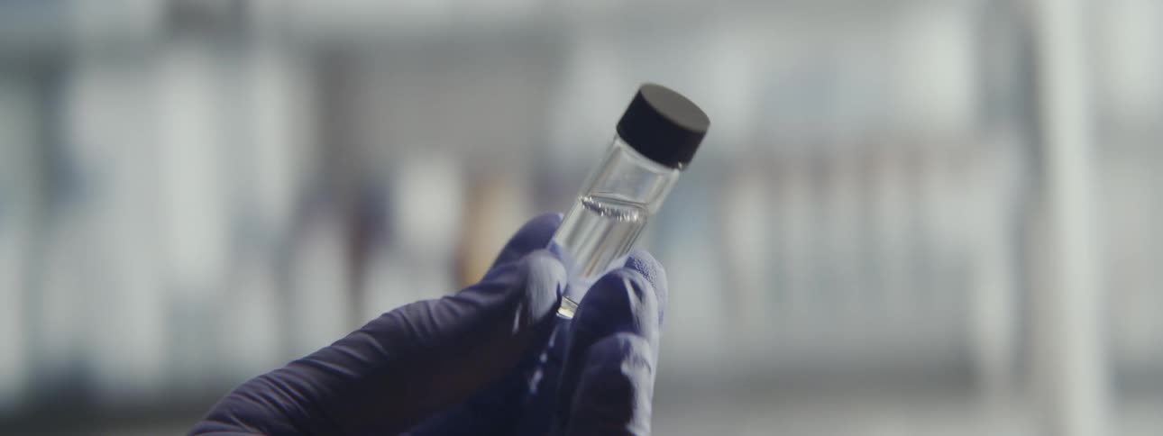 A scientist in purple gloves examines a small glass vial containing a clear liquid in a laboratory