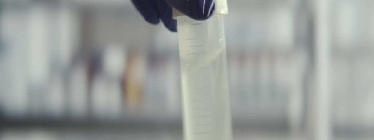 Close-up of a scientist in blue gloves swirling a clear liquid sample in a test tube inside a laboratory